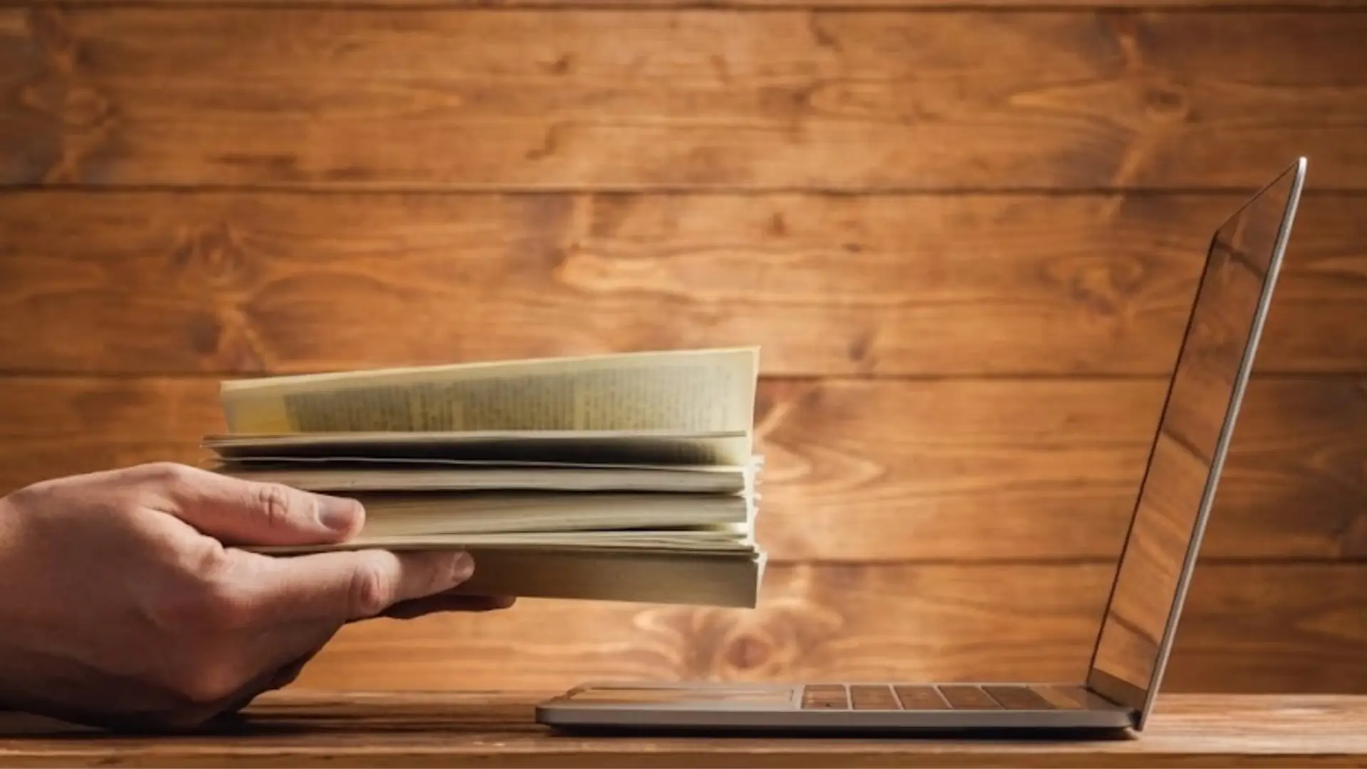 Hand holding stack of books or papers in front of open laptop on wooden desk, symbolizing intersection of traditional learning and digital technology.