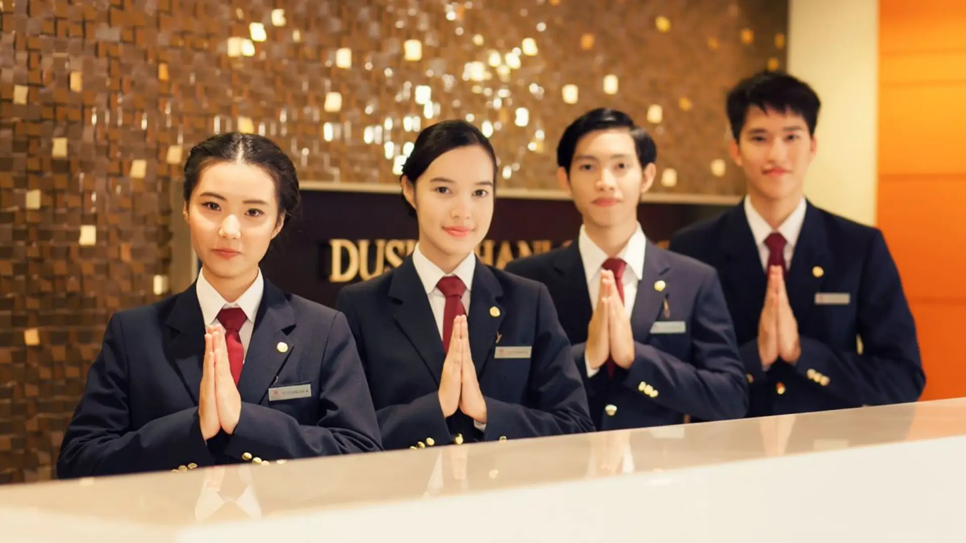 Four hotel staff in formal uniforms performing Thai “wai” greeting gesture behind reception desk, decorative wall with Dusit Thani branding, symbolizing hospitality and cultural respect.