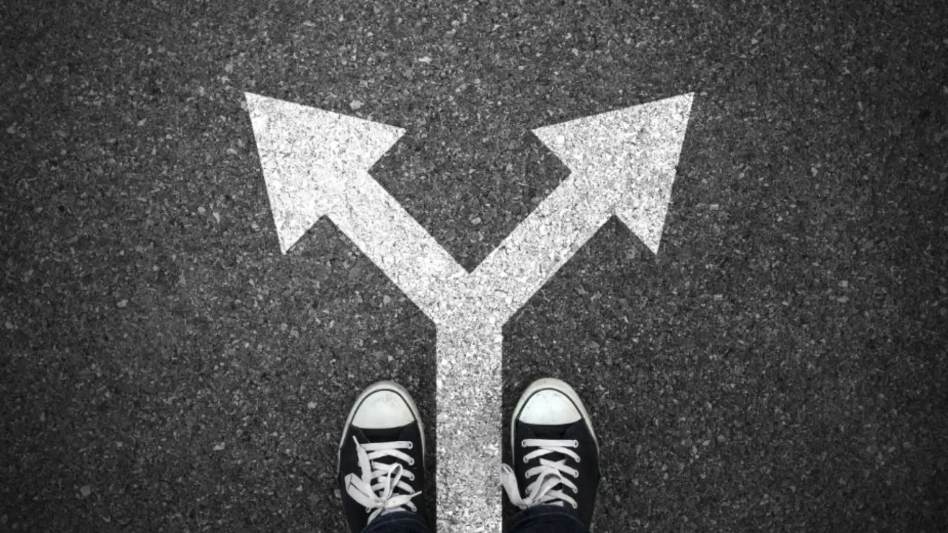 Feet in sneakers standing at fork in road with two white arrows pointing left and right, symbolizing choice and decision‑making.