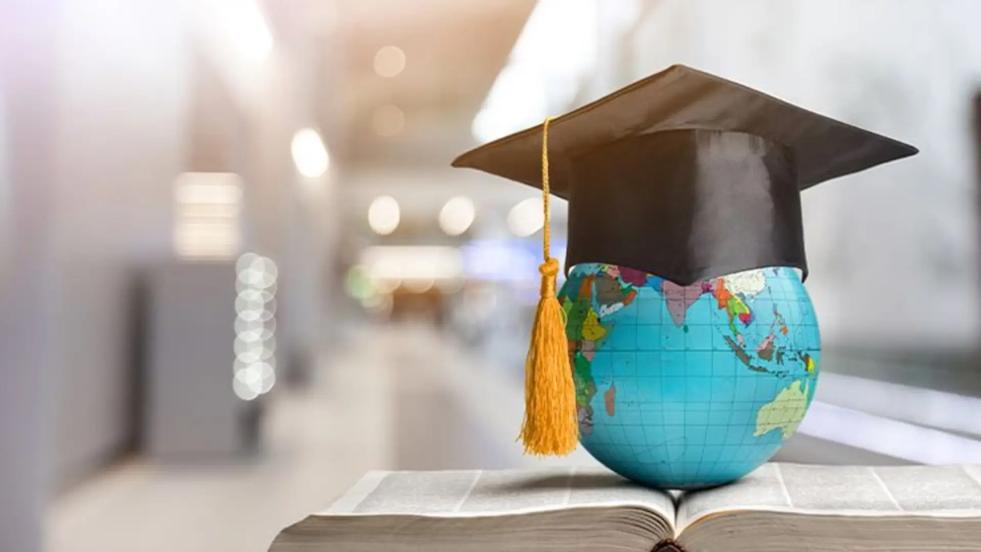Small globe wearing black graduation cap with yellow tassel placed on open book, symbolizing global education and academic achievement.