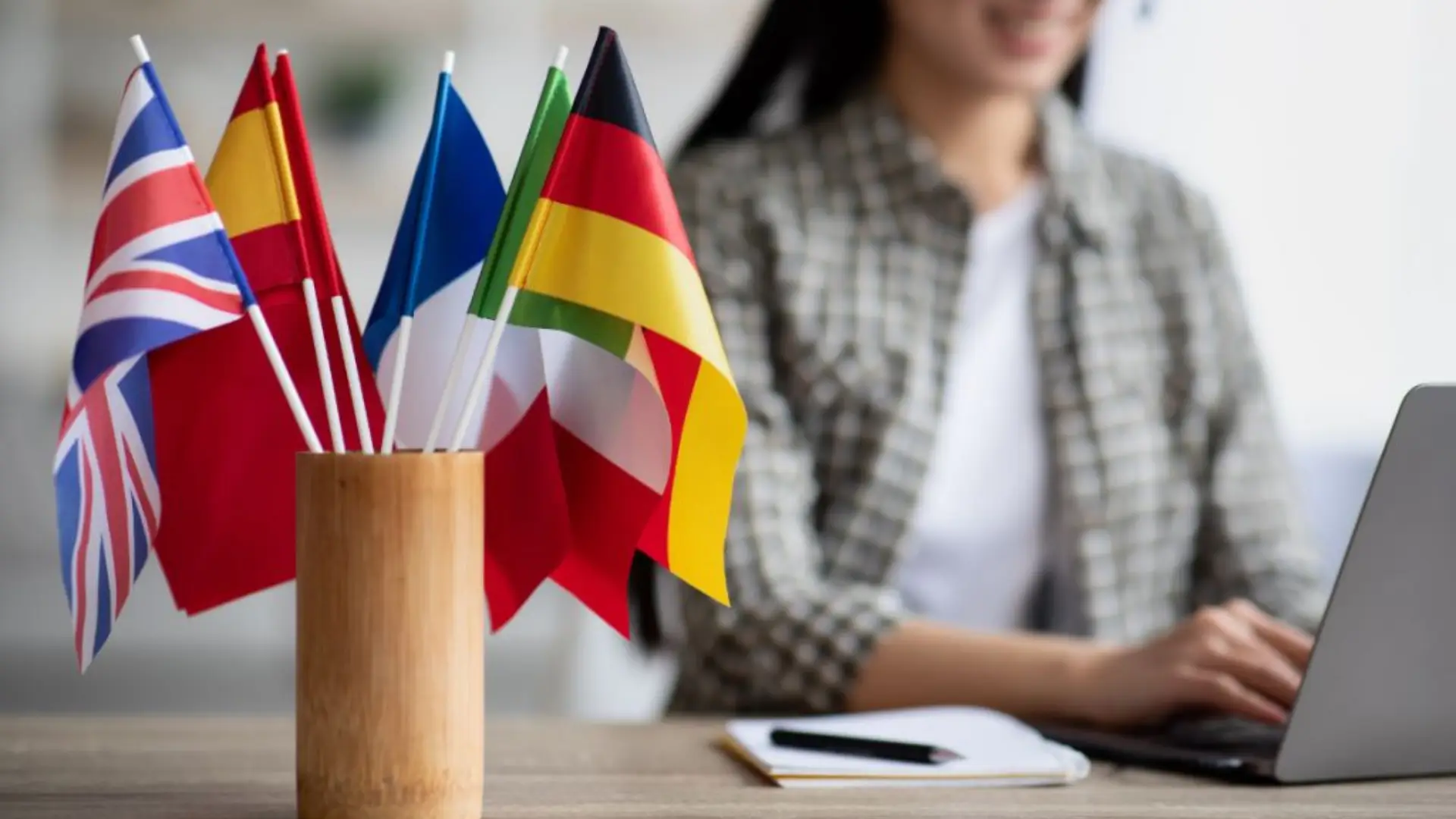 Wooden container holding small flags of UK, Spain, France, Finland, Japan, Germany, and Italy on desk with person typing on laptop in background, symbolizing multilingual collaboration.